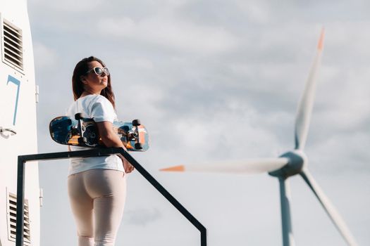 A girl in white clothes and glasses with a skate in her hands is photographed near large wind turbines in a field with trees.Modern woman with a riding Board in a field with windmills.