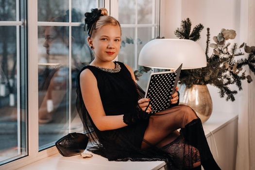 A stylish girl in a black dress sits on the windowsill by the window with a book in her hands.