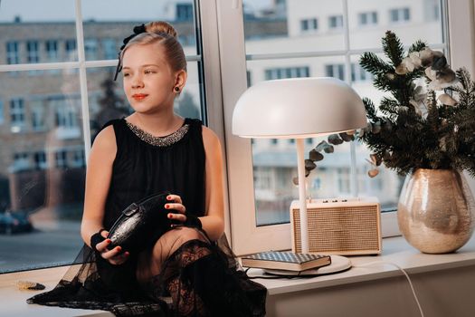 A stylish girl in a black dress sits on the windowsill by the window with a purse in her hands.