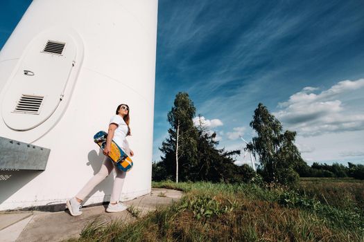 A girl in white clothes and glasses with a skate in her hands is photographed near large wind turbines in a field with trees.Modern woman with a riding Board in a field with windmills.