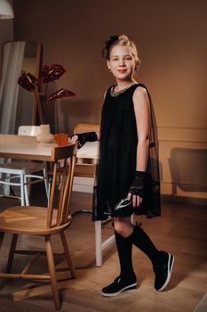 A stylish little girl in a black dress stands in the interior near the table.