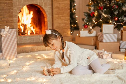 Kid playing with toy on Christmas morning, sitting on carpet in decorated living room near x-mas tree and fireplace, smiling happy little girl with pigtail holding tiny toys in hands, new year.