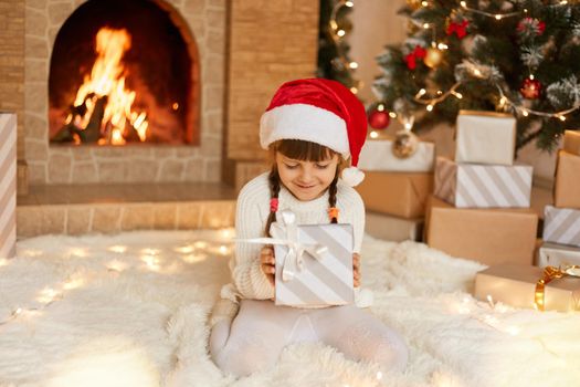 Cute little child in red festive hat with Christmas gift box posing at home while sitting on floor on soft carpet near fireplace and xmas tree. Little charming girl with pigtails holds gift box.