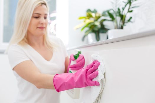 Woman in the kitchen is smiling and wiping dust using a spray and a duster while cleaning her house, close-up