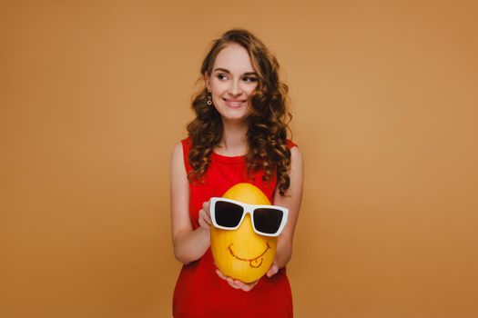 beautiful young girl holds hands melon. The melon is wearing glasses and an emoticon is drawn. Girl in a red dress.