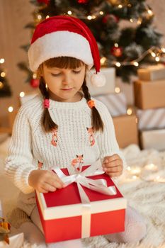 Little european female kid with two pigtails sitting on floor on background of present boxes and lights, holding gift box in hands and looking on it, keeping fingers on ribbon.