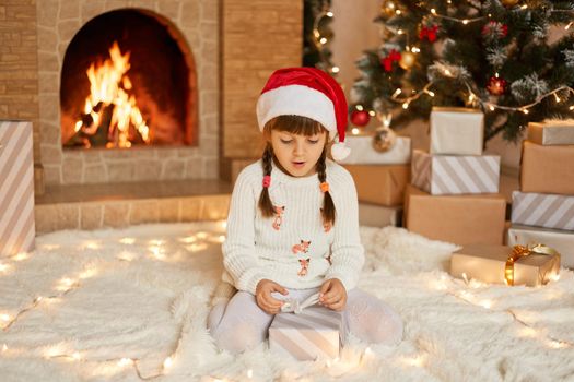 Astonished little girl on Christmas eve at fireplace. Kid opens Xmas present. Child in decorated living room with traditional fire place. Cozy winter evening at home. Surprised female kid on floor.