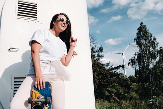 A girl in white clothes and glasses with a skate in her hands is photographed near large wind turbines in a field with trees.Modern woman with a riding Board in a field with windmills.