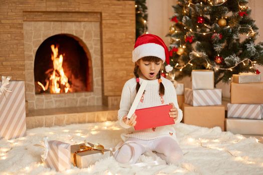 Little girl's excitement from opening presents on Christmas, female child sitting on floor in santa hat, keeps mouth opened, being surprised and astonished, posing in living room with xmas decoration.