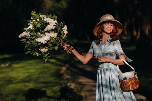 Portrait of a beautiful girl with long hair, a straw hat and a long summer dress with lilac flowers in the garden.