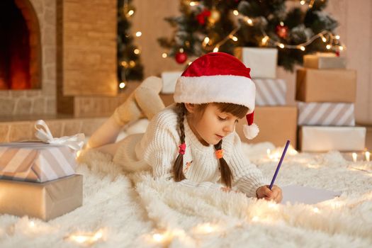 Pretty girl in xmas hat writes letter to Santa while laying on floor on white carpet, female child with two pigtails posing near christmas tree and gift boxes with pen or pencil in hand.