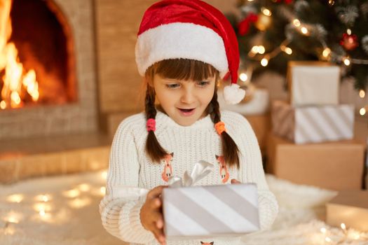 Cute female kid holding christmas gift. Little child having fun near Christmas tree in living room. Surprised child wearing santa hat and white sweater, looking at present box with opened mouth.
