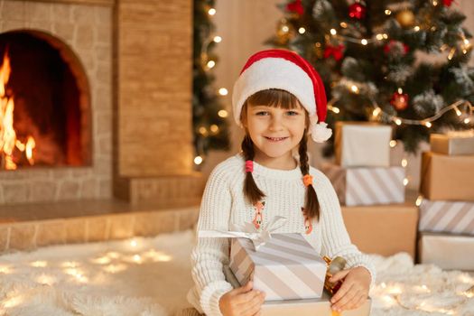 Beautiful smiling little girl with gifts sitting on floor and looking at camera, wearing white jumper and red hat, child posing near fireplace and Christmas tree.