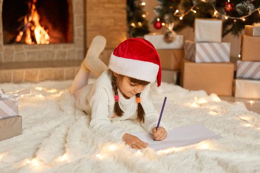 Happy girl in red Christmas hat writing letter to Santa Claus or drawing picture, female child wearing white sweater and warm socks, kid posing in festive living room with fireplace.