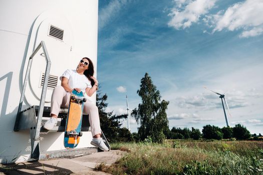 A girl in white clothes with a skate in her hands is photographed near large wind turbines in a field with trees.Modern woman with a Board for riding in a field with windmills.