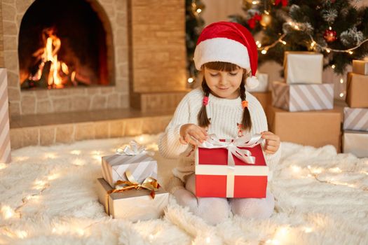 Little girl opening christmas present, keeping fingers on ribbons, looking at gift box with smile, child wearing santa claus hat posing on floor near fireplace and xmas tree.