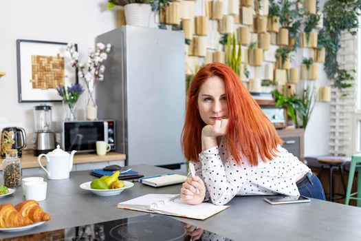 smiling attractive woman with red hair sits with notepad and pen in the kitchen at home. maternity education, distance learning and home office