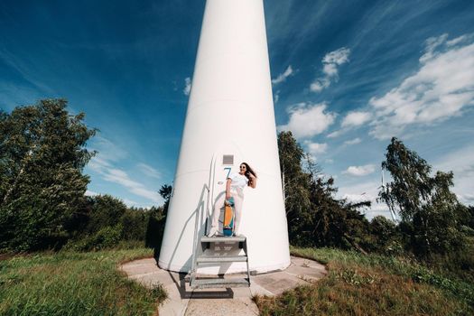 A girl in white clothes and glasses with a skate in her hands is photographed near large wind turbines in a field with trees.Modern woman with a riding Board in a field with windmills.