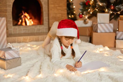 Little girl in red christmas hat writes letter to Santa Claus, adorable child lying on floor near fireplace, wearing white warm jumper, lying on floor on soft carpet.