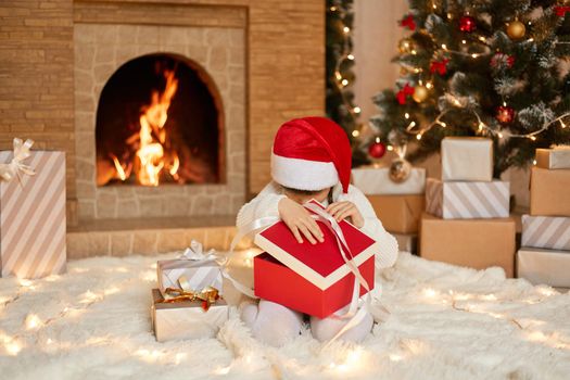 Little girl in santa hat opening gift box, looking inside it, wearing white jumper and red santa claus hat, sitting on floor in festive living room, posing with fireplace and xmas tree on background.