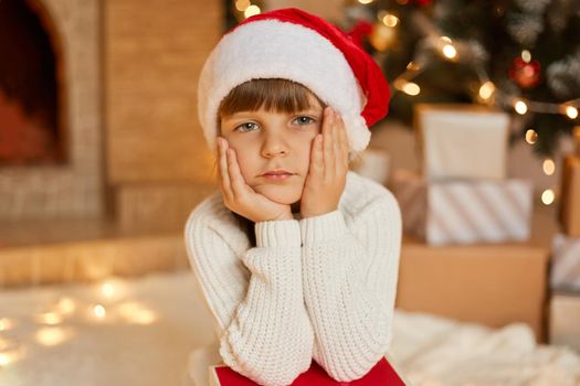 Little bored girl waiting for Christmas, sitting in festive living room, keeping hands on cheek, looks at camera with sad expression, wearing white sweater and red santa hat.