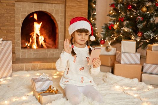 Kid in santa hat having web chat call with somebody, looking at screen and waving hand. Cute preschooler girl with two pigtails wearing santa claus hat and white pullover.
