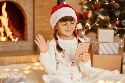 Christmas online, congratulations from home, smiling little girl using smart phone for video call. Child talks to friends and parents, waves hands to greet, wearing santa hat, poses in festive room.