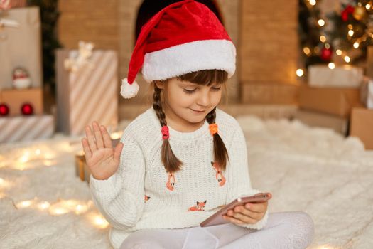 Little cute female kid with pigtails congratulation from home, smiling little girl using smart phone for video call, talks to somebody, waves hands to greet, wearing santa hat and white sweater.