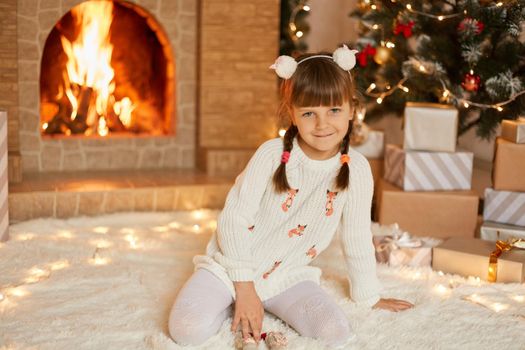 Xmas girl sitting next to festive tree and looking directly at camera, child looking directly at camera, wearing white sweater and tights, happy kid celebrating new year at home near fireplace.