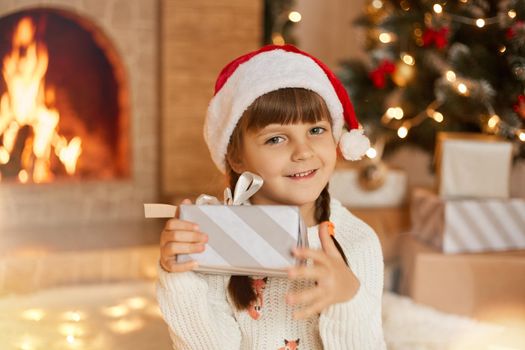 Happy child girl with Christmas present looking directly at camera, wearing red santa claus hat, holding gift box with ribbon in hands, posing at home with fireplace and x-mas tree on background.