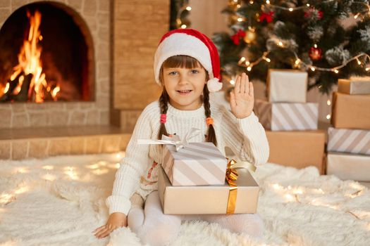 happy child girl sitting near Christmas tree on Christmas Eve on floor and waving hand to camera, posing with stack of gift boxes, kid wearing casual clothing and red santa hat.