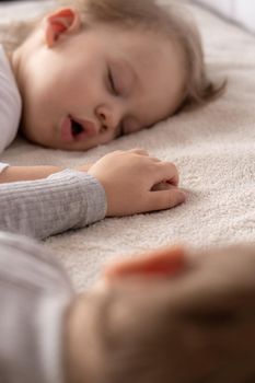 Childhood, sleep, relaxation, family, lifestyle concept - two young children 2 and 3 years old dressed in white and beige bodysuit sleep on a beige and white bed at lunch holding hands top view