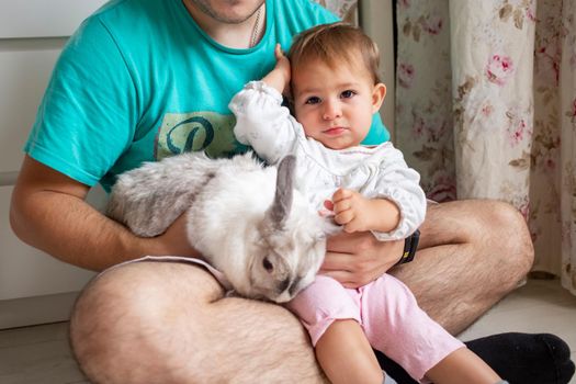 adorable baby sits in dad's arms and strokes a decorative rabbit. father shows little child easter bunny. domestic animals in a family with children