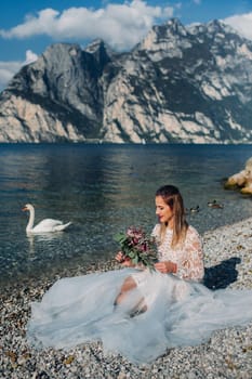 a girl in a smart white dress is sitting on the embankment of lake Garda.A woman is photographed against the background of a mountain and lake in Italy.Torbole.