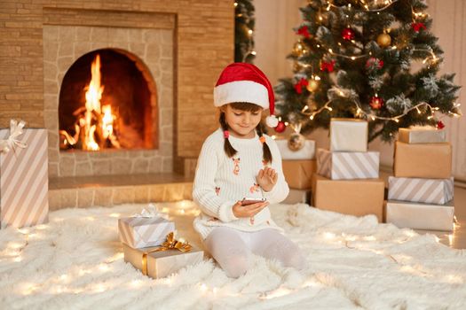 Christmas mood. Cute little child holding phone in hands, get smart phone as xmas present, sitting on floor on soft carpet near gift boxes and fireplace, dresses festive santa claus hat.