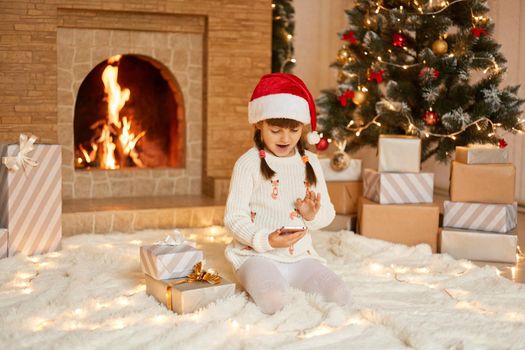 Little girl with pigtails communicates with relatives on phone and thanks them for gifts, waving hand to smartphone's camera, saying hello, wearing white sweater and santa claus hat.