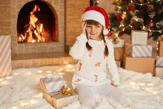 Happy little girl in beautiful white sweater and red santa hat in front of New Year Christmas tree, smiling, poses and holding smart phone near ear, sits near gift boxes, talking and thanks for gifts