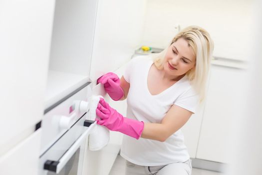 Woman in the kitchen is smiling and wiping dust using a spray and a duster while cleaning her house, close-up