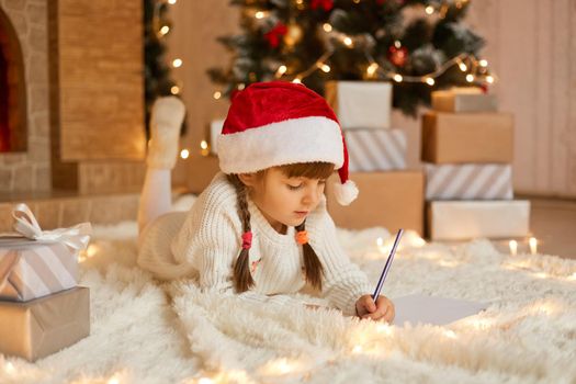 Cute child girl writing letter santa at home near gift boxes while lying on floor on soft carpet, kid with pigtails drawing picture for present, poses near xmas tree.