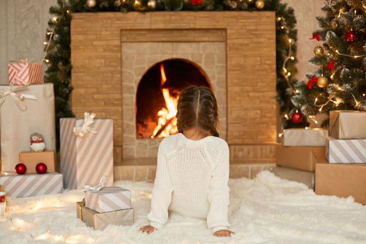 Little girl sitting by fireplace on Christmas eve, looking at fire while sitting on soft carpet, waiting new Year Eve, wants to open her present boxes.