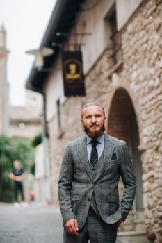 A man with a beard in a strict grey three-piece suit with a tie in the old town of Sirmione, a Stylish man in a grey suit in Italy.