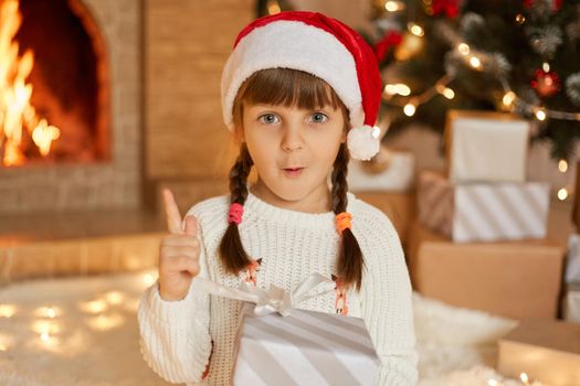 Little Santa girl in red hat and white jumper, posing indoor in festive room, having surprised facial expression, having great idea, pointing finger up, wants to give present for somebody.