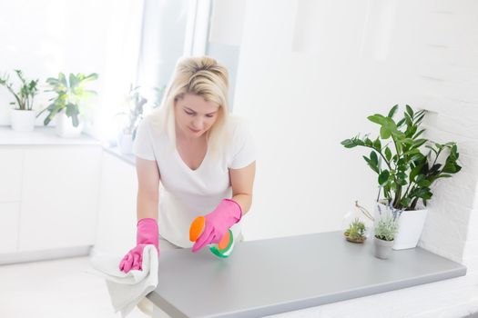 Woman in the kitchen is smiling and wiping dust using a spray and a duster while cleaning her house, close-up