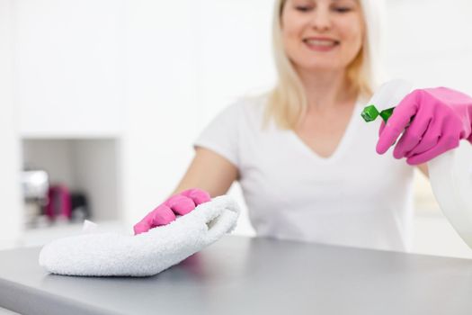 Woman in the kitchen is smiling and wiping dust using a spray and a duster while cleaning her house, close-up