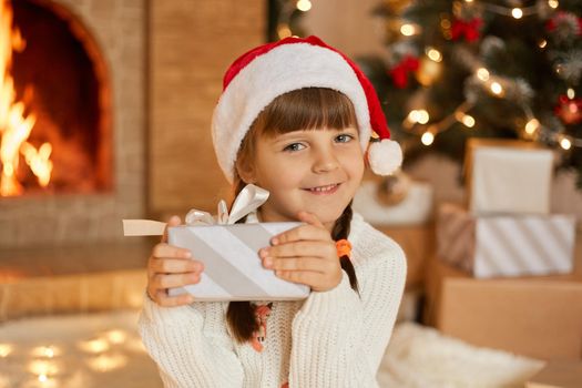 Happy little girl with present at Christmas Eve, sitting near xmas tree and fireplace, charming smiling child looking at camera and holding striped gift box with bow.