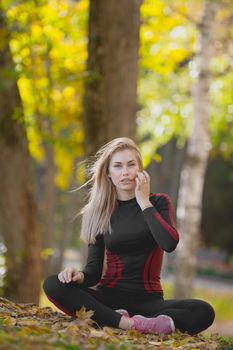 Young attractive fitness female sitting cross-legged at workout in autumn park, straightens her waving in the wind hair, telephoto