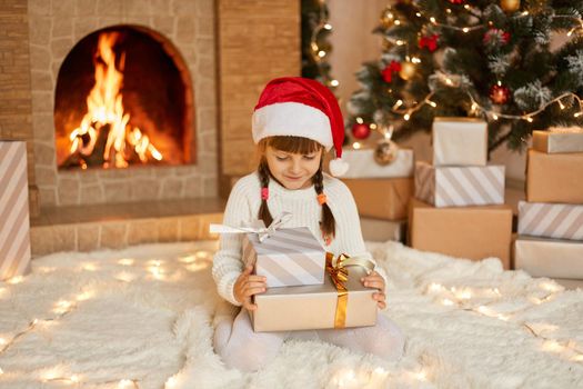 Happy little girl in Santa hat holding Christmas gift boxes while sitting on floor, child wearing white jumper and red santa claus, posing in festive room near xmas tree and fireplace.