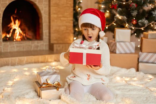 Charming adorable female kid with present box in hand sitting on floor, looking at her gift with surprised facial expression, keeps mouth widely opened, wearing santa hat, pose i festive living room.