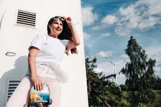 A girl in white clothes and glasses with a skate in her hands is photographed near large wind turbines in a field with trees.Modern woman with a riding Board in a field with windmills.