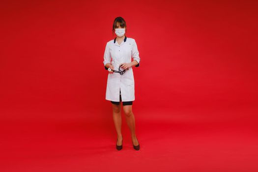 A young doctor in a white coat and medical mask stands on a red background.